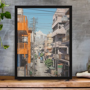 Op de afbeelding: Een ingelijste illustratie van een smalle straat in een Japanse stad. De straat is omzoomd met gebouwen, waarvan sommige oranje en andere lichtroze zijn geverfd. De straat is omzoomd met stroomleidingen en er zijn bomen en struiken aan beide kanten. Er rijdt een kleine vrachtwagen over de straat.