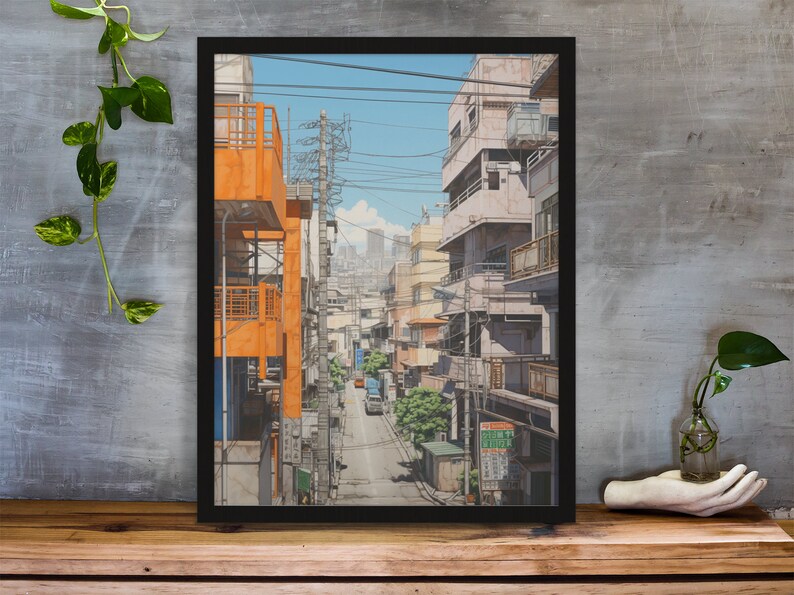 Op de afbeelding: Een ingelijste illustratie van een smalle straat in een Japanse stad. De straat is omzoomd met gebouwen, waarvan sommige oranje en andere lichtroze zijn geverfd. De straat is omzoomd met stroomleidingen en er zijn bomen en struiken aan beide kanten. Er rijdt een kleine vrachtwagen over de straat.
