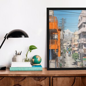 Op de afbeelding: Een ingelijst afdruk van een kleurrijke illustratie van een smalle straat in een stad met gebouwen aan beide kanten. De gebouwen zijn een mix van oranje, wit en grijs. De straat is bezaaid met stroomleidingen en er rijden auto's over de straat.