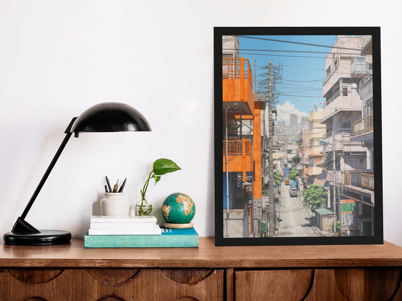 Op de afbeelding: Een ingelijst afdruk van een kleurrijke illustratie van een smalle straat in een stad met gebouwen aan beide kanten. De gebouwen zijn een mix van oranje, wit en grijs. De straat is bezaaid met stroomleidingen en er rijden auto's over de straat.