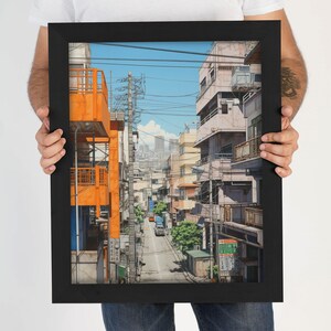 Op de afbeelding: Een ingelijste illustratie van een smalle straat in een Japanse stad. De straat is omzoomd met gebouwen, waarvan sommige oranje geverfd zijn. Er lopen bovengrondse stroomleidingen en een auto rijdt over de straat.