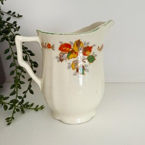 May include: Cream ceramic pitcher with a handle and spout, decorated with a floral pattern in red, yellow, and green. The rim has a green trim. A vintage-style decorative piece. The pitcher is approximately 15 cm tall.
