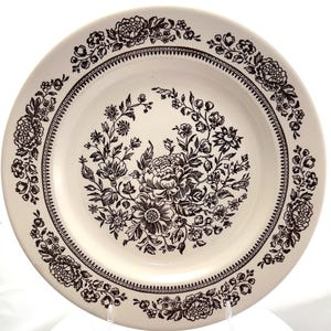 May include: A round ceramic plate with an off-white base color featuring a dark brown floral pattern. The center of the plate displays a detailed bouquet of flowers, while the rim is decorated with a repeating floral border and a geometric band.