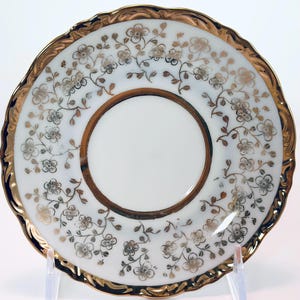 May include: A white saucer with a gold rim and a gold floral pattern. The center of the saucer is white, and the rim has a scalloped edge. The floral design includes small flowers and leaves.