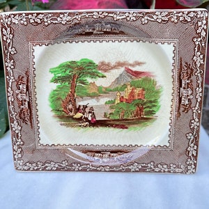 May include: A square ceramic dish with a brown and white border. The center of the dish features a colorful landscape scene with a castle, trees, and a lake.