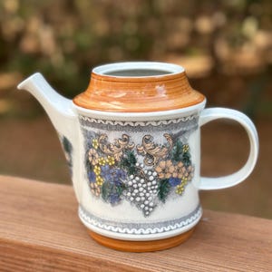 May include: A white ceramic teapot with a decorative grape and floral design. The teapot has a brown lid and base, a curved spout, and a rounded handle. The design includes blue, yellow, and peach-colored flowers and green leaves.