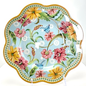 May include: A decorative ceramic plate with a scalloped edge. The plate features a light blue background with a floral design of pink, yellow, and white flowers, green leaves, and blue butterflies. The rim is gold with a green and white border.