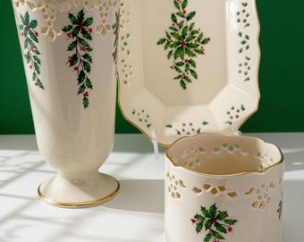 May include: A cream-coloured ceramic set featuring a tall vase, a rectangular tray, and a short cylindrical candle holder. Each piece has gold-trimmed edges, intricate cut-out lattice patterns, and a painted green holly and red berry motif.