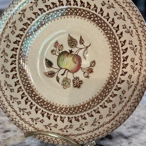 May include: A vintage ceramic plate with a cream-colored background and a brown floral design. The plate features an apple motif in the center, surrounded by intricate patterns and a scalloped edge. The plate is displayed on a silver metal stand.