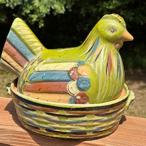 May include: A hand-painted ceramic chicken figurine in shades of green, yellow, blue, and brown. The chicken sits atop a matching basket-like base. The figurine has a detailed design, with a textured appearance and a rustic aesthetic.