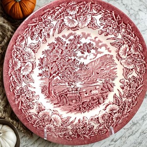 May include: A round ceramic plate featuring a deep red transferware pattern. The center depicts a rural landscape with a horse-drawn cart, sheep, and trees. The wide border is decorated with detailed floral motifs including blossoms and leaves.