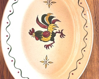 May include: A cream-colored oval serving dish with a green and red border. The center of the dish features a hand-painted rooster with yellow, green, and red accents. The rooster is surrounded by two yellow four-pointed stars.