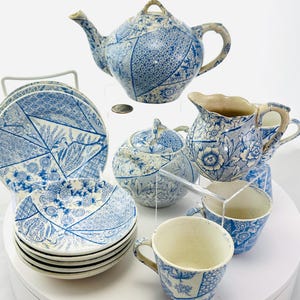May include: A vintage blue and white porcelain tea set. The set includes a teapot, creamer, sugar bowl, cups, and saucers. The pieces feature a patchwork design with floral and geometric patterns. The tea set is displayed on a white surface.