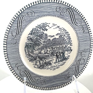May include: A vintage decorative plate with a blue and white pastoral scene. The plate features a detailed landscape illustration in the center, surrounded by a blue patterned border and decorative script. The plate is displayed on a clear stand.