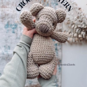 May include: A crocheted teddy bear in a light brown color, held up in front of a soft background. The bear has a textured appearance, with visible stitches and a detailed snout. The text "CROCHET PATTERN" is arched above the bear.