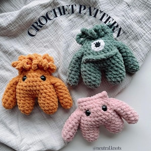Peut inclure: Trois jouets monstres crochetés à la main en orange, vert et rose. Chacun a un corps rond, quatre membres trapus et deux yeux noirs en forme de bouton. Le texte "CROCHET PATTERN" est arqué au-dessus des jouets.