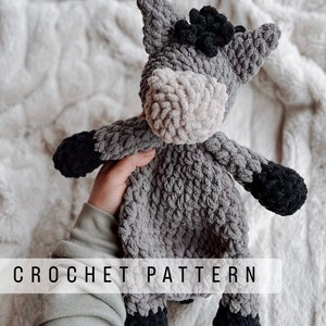 May include: A gray and black crocheted donkey toy with a textured surface. The toy is being held by a hand. CROCHET PATTERN is written in white text at the bottom of the image.