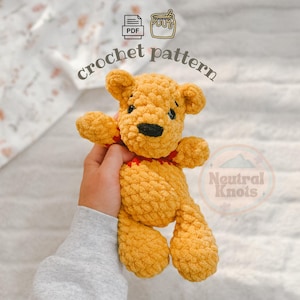 May include: A golden yellow crochet bear toy with a black nose and a red collar. The image includes the text "crochet pattern" and "Neutral Knots" with a PDF icon and a honey pot icon. The bear has a textured, plush appearance with small ears.