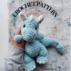 May include: A crocheted blue and white dinosaur toy. The plush toy has a rounded body, small arms and legs, and a long tail. The text "CROCHET PATTERN" is arched above the toy.