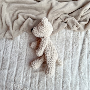 May include: A white crocheted teddy bear lying on a white fluffy blanket. The bear is curled up in a sleeping position.