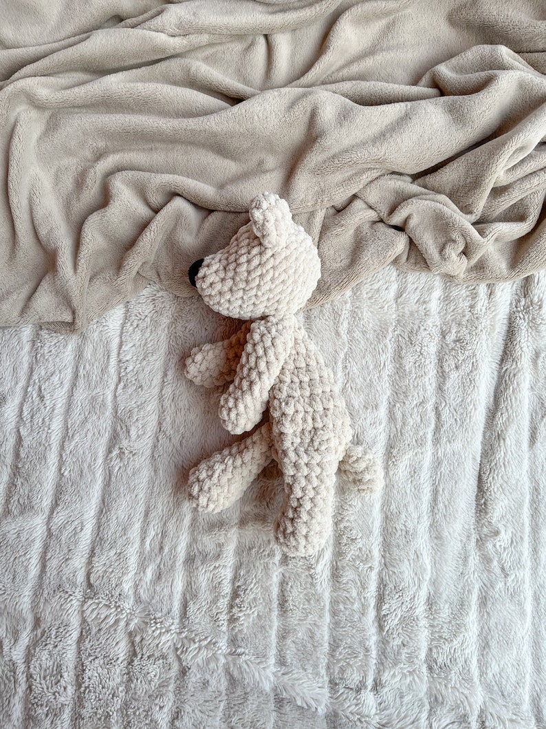 May include: A white crocheted teddy bear lying on a white fluffy blanket. The bear is curled up in a sleeping position.