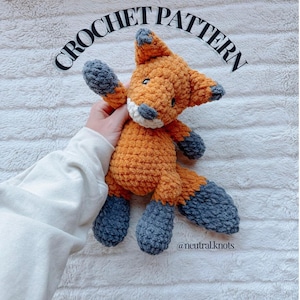May include: A plush crochet fox toy with orange fur, dark gray paws, ears, and tail tip, and a white muzzle. The text CROCHET PATTERN is printed in black above the fox, and the text @neutral.knots is printed in black below the fox.