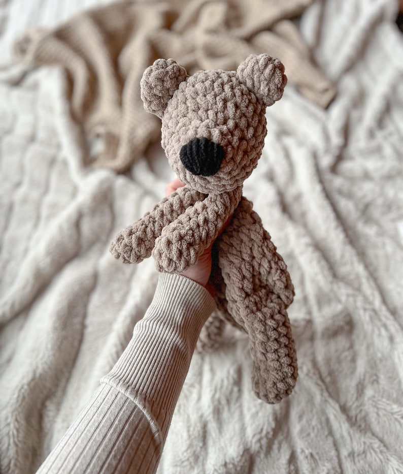 May include: A crocheted brown teddy bear with black eyes and a nose. The bear is being held by a hand wearing a beige sweater.