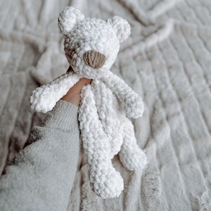 May include: A white crocheted teddy bear with a brown nose. The bear is being held by a hand wearing a white sweater.