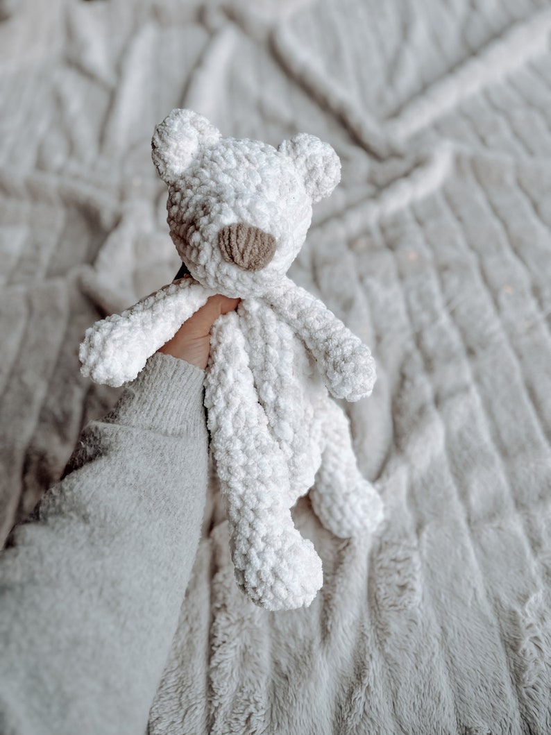 May include: A white crocheted teddy bear with a brown nose. The bear is being held by a hand wearing a white sweater.