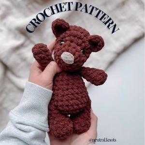 May include: A handmade, dark brown crochet bear with a light beige snout and small black eyes. The image includes the text "CROCHET PATTERN" in a curved font. The bear is held in hands, suggesting a soft, plush toy.