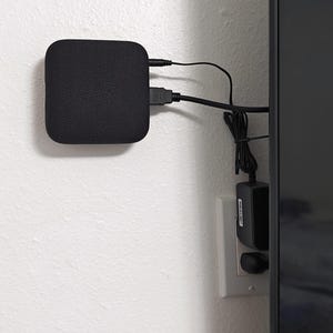 May include: A black square-shaped speaker with a fabric grille is mounted on a white wall. The speaker is connected to a black television with a black HDMI cable and a black power cord. The power cord is plugged into a white electrical outlet.