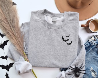 Peut inclure: Sweat-shirt gris avec un motif de chauve-souris noir sur le devant. Le sweat-shirt est posé à plat sur une surface blanche avec un chapeau marron, une tasse de café et un jean.