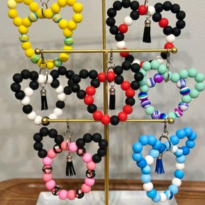 May include: A display of beaded heart-shaped keychains in various colors, including yellow, black, red, pink, and blue. Each keychain features a tassel and a metal clasp. The keychains are arranged on a gold-colored metal stand.