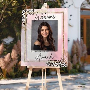 Puede incluir: Un letrero de bienvenida rosa y dorado sobre un caballete. El letrero presenta una foto de una mujer y el texto "Bienvenida a la fiesta de graduación de Amanda". El letrero tiene un borde con estampado de leopardo y detalles dorados.