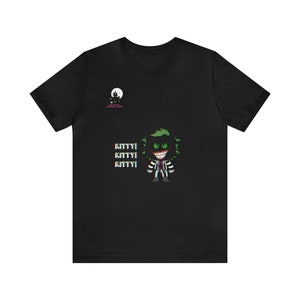 Op de afbeelding: Zwart t-shirt met een cartoonfiguur met groen haar, groene ogen en een gestreept pak. Het woord "KITTY!" wordt driemaal herhaald. Er is ook een kleine afbeelding van een spookhuis en een maan.