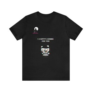 Op de afbeelding: Zwart t-shirt met een afbeelding van Hello Kitty verkleed als Freddy Krueger, met een mes. De tekst "1 2 Kitty's Coming For You" staat op het shirt, samen met een kleine afbeelding van een spookhuis.