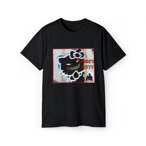 Op de afbeelding: Zwart t-shirt met een vervormd beeld van een cartoonkat met de tekst "HERE'S KITTY". De afbeelding heeft een rode rand en een spookhuislogo. Het ontwerp heeft een 3D-effect.