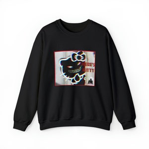 Op de afbeelding: Zwarte crewneck sweatshirt met een afbeelding van een vervormd Hello Kitty-achtig karakter met scherpe tanden en gloeiende ogen. De afbeelding bevat de tekst "HERE'S KITTY" in het rood, tegen een houten achtergrond.