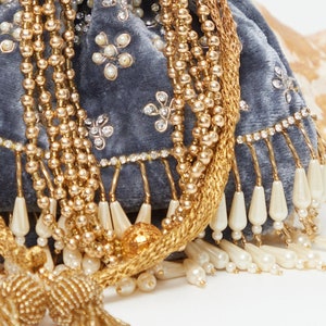 K&ouml;nnte beinhalten: Eine graue Samt-Kordeltasche mit goldenen und wei&szlig;en Perlenverzierungen. Die Tasche hat einen Quasten und einen Fransen aus wei&szlig;en Perlen.