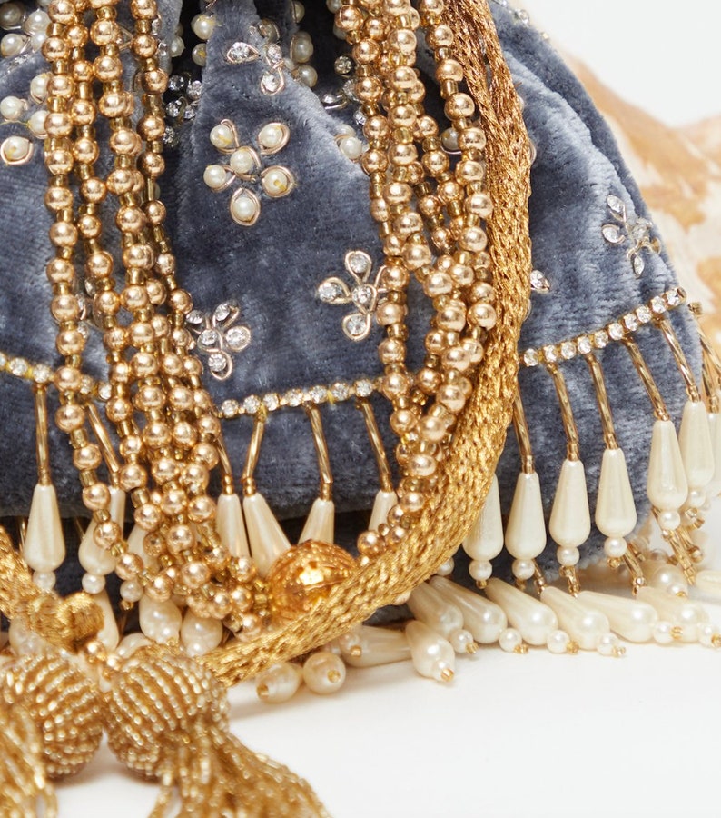 K&ouml;nnte beinhalten: Eine graue Samt-Kordeltasche mit goldenen und wei&szlig;en Perlenverzierungen. Die Tasche hat einen Quasten und einen Fransen aus wei&szlig;en Perlen.