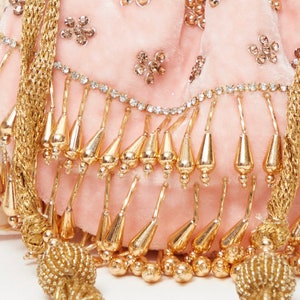 K&ouml;nnte beinhalten: Eine rosa Samt-Kordeltasche mit goldenen und silbernen Verzierungen. Die Tasche hat einen Fransen aus goldenen Perlen und einen goldenen Quasten.