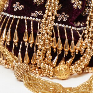 K&ouml;nnte beinhalten: Nahaufnahme einer bordeauxroten Samt-Tasche mit goldenen Perlenverzierungen und einem Quastenanh&auml;nger. Die Tasche ist mit Strasssteinen verziert und hat einen Fransenrand.