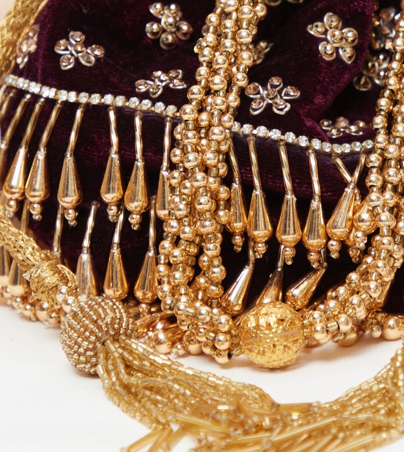 K&ouml;nnte beinhalten: Nahaufnahme einer bordeauxroten Samt-Tasche mit goldenen Perlenverzierungen und einem Quastenanh&auml;nger. Die Tasche ist mit Strasssteinen verziert und hat einen Fransenrand.