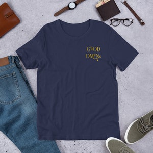 May include: A navy blue t-shirt with a gold embroidered design that says "GOOD OMENS" with a small devil tail.
