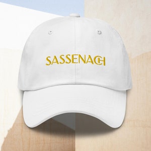 May include: White baseball cap with a gold embroidered word "Sassenach" on the front.