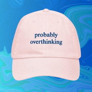 May include: A light pink baseball cap with the embroidered text "probably overthinking" in navy blue. The cap has a curved brim and a rounded top. The background is a blue and teal abstract design.