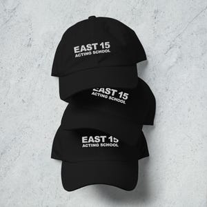 Peut inclure: Quatre casquettes de baseball noires empilées. Chaque casquette porte l'inscription blanche "EAST 15 ACTING SCHOOL". Les casquettes sont disposées verticalement, les visières orientées vers l'avant.