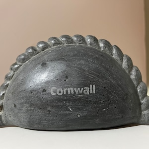 Könnte beinhalten: Eine graue, steinartige Skulptur in Halbmondform mit einem seilartigen Rand. Die Skulptur trägt die Aufschrift "Cornwall" auf der Vorderseite.