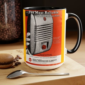 May include: A white ceramic mug with a black rim and handle. The mug features a vintage advertisement for RCA Drive-In Speakers. The ad features a black and white image of a speaker with the text "The Most Reliable of Drive-In Speakers" and "RCA Radio Corporation of America".