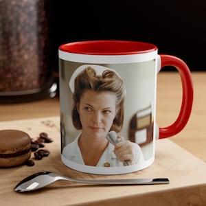 May include: A white ceramic mug with a red rim and handle. The mug features a color photograph of a woman wearing a white nurse's uniform and cap. She is holding a medical device in her right hand and looking directly at the camera.
